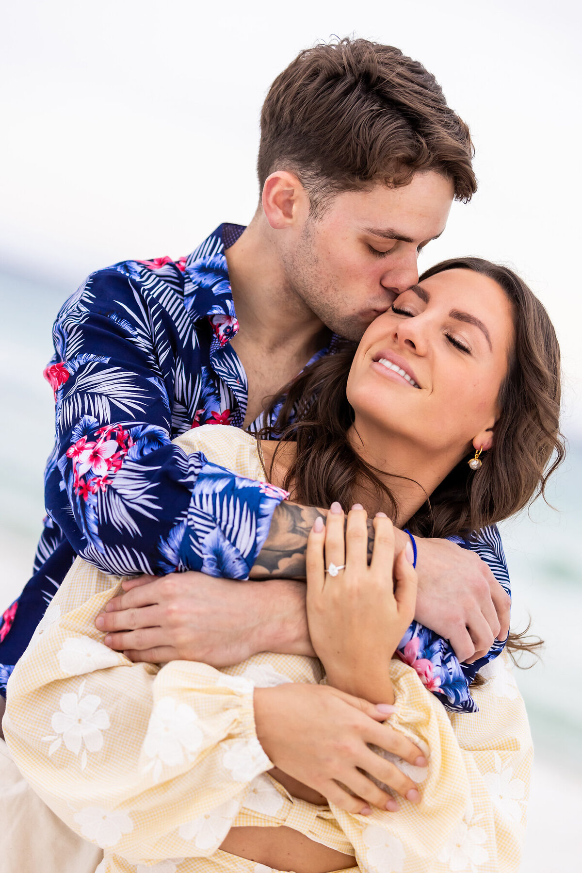 Happy couple that are newly engaged sitting in sand hugging while engagement ring is visible.