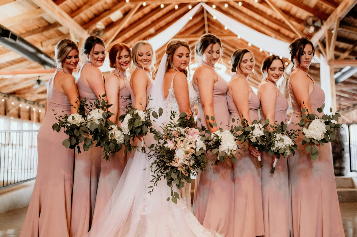 bride and her bridesmaids standing in a line while holding hteir bouquets down to their sides while looking down their arms to their florals