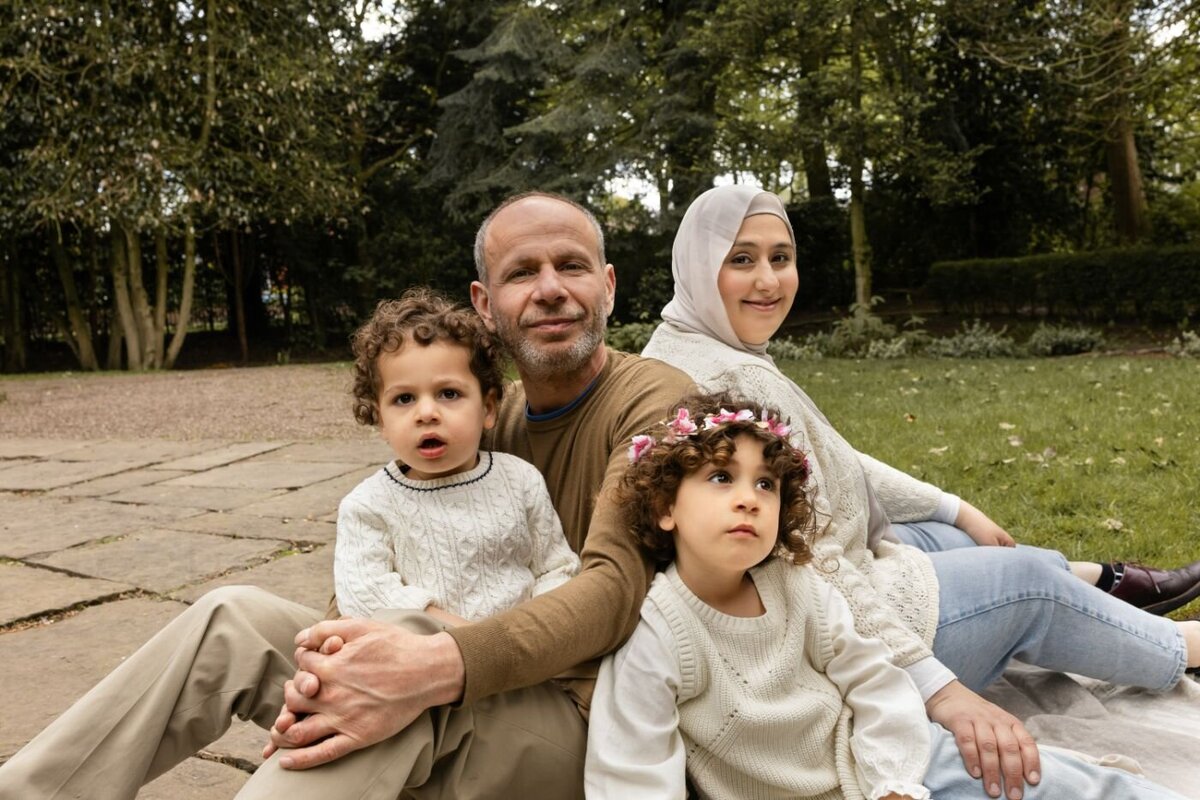 A smiling family of four, including parents and two young children with curly hair (one wearing a floral crown and the mother in a hijab), sit together on a stone surface outdoors