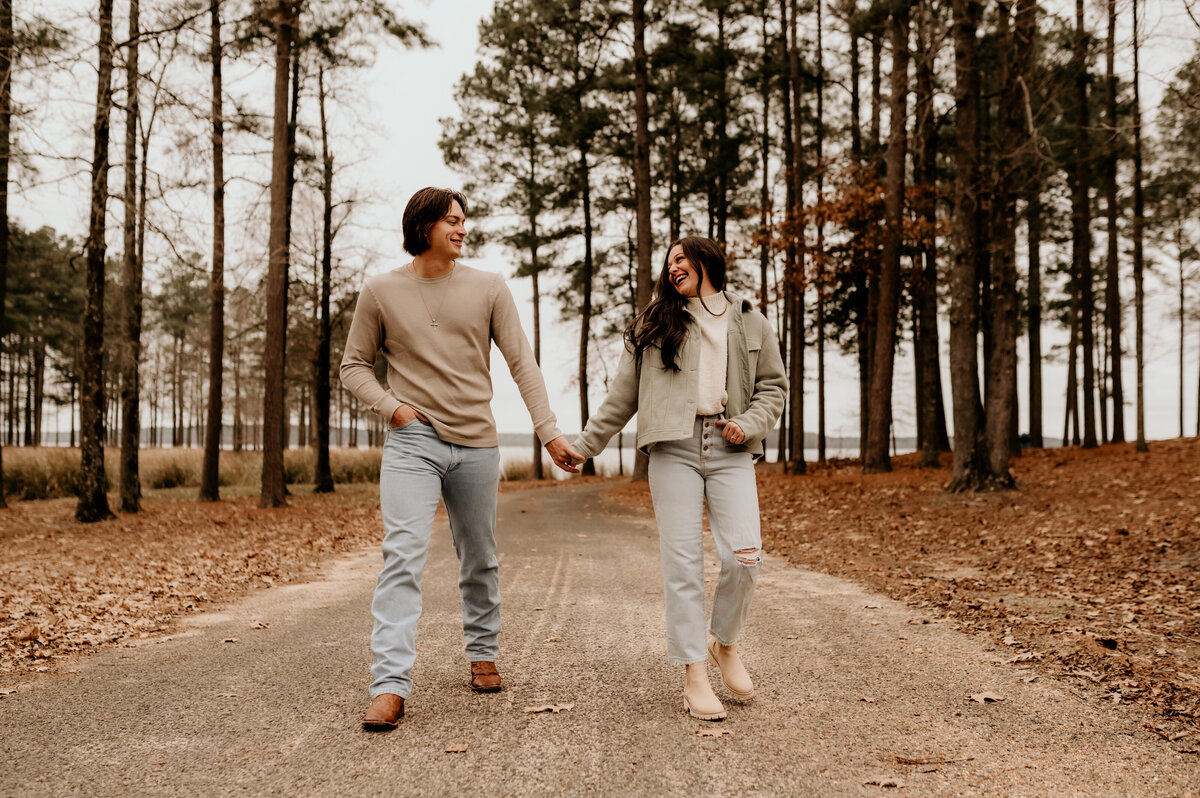 Little rock engagement photographer captured man and woman holding hands and running to the camera with the forest behind them as they look at one another and smile