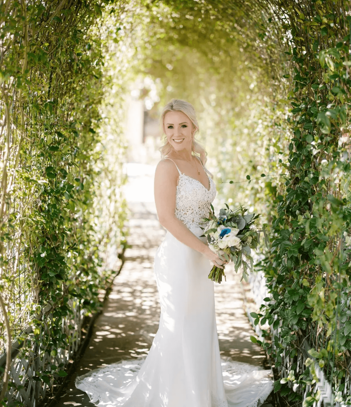 Bride posing in a sunlit garden archway in Dallas, showcasing professional bridal makeup, soft updo styling, and natural wedding glow