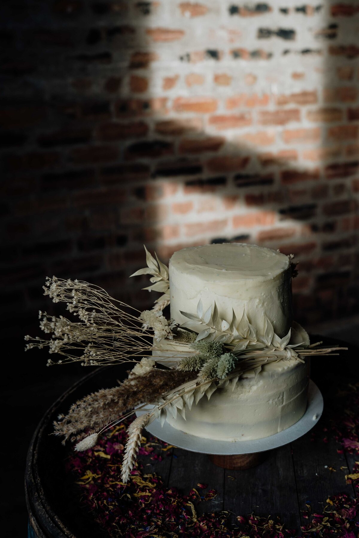 A two-tier white wedding cake, adorned with dried flowers and leaves, sits elegantly on a wooden surface. The cake is photographed in front of a brick wall background, which casts a shadow over the cake, enhancing its rustic and moody charm.