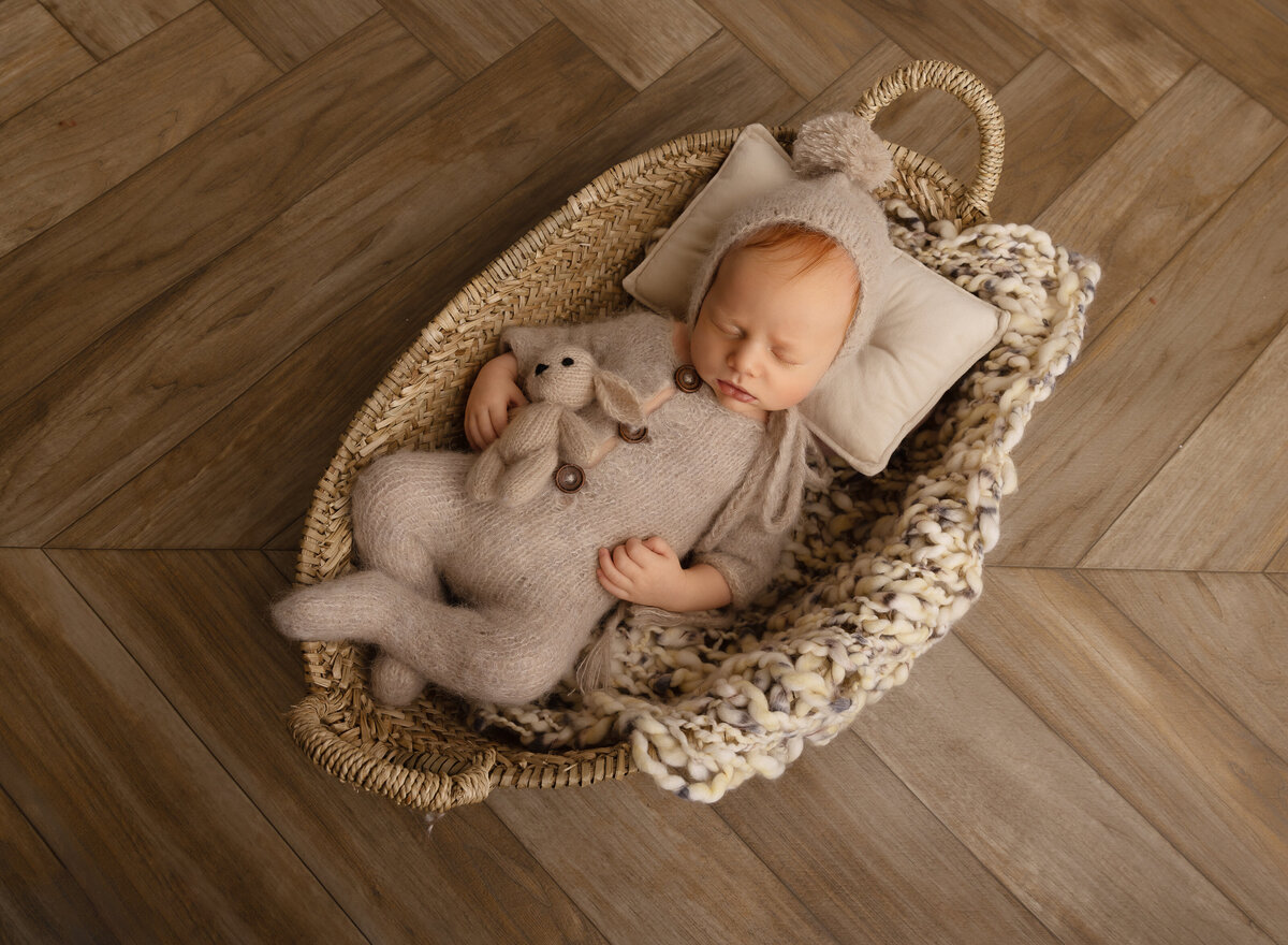 Bay in beige onesie is in a moses basket holding a felt teddy for his newborn photoshoot. Captured by top Brooklyn newborn photographer Chaya Bornstein
