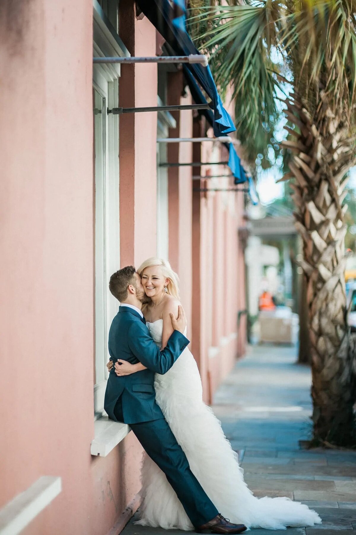 A groom leaning against a building hugging a bride.