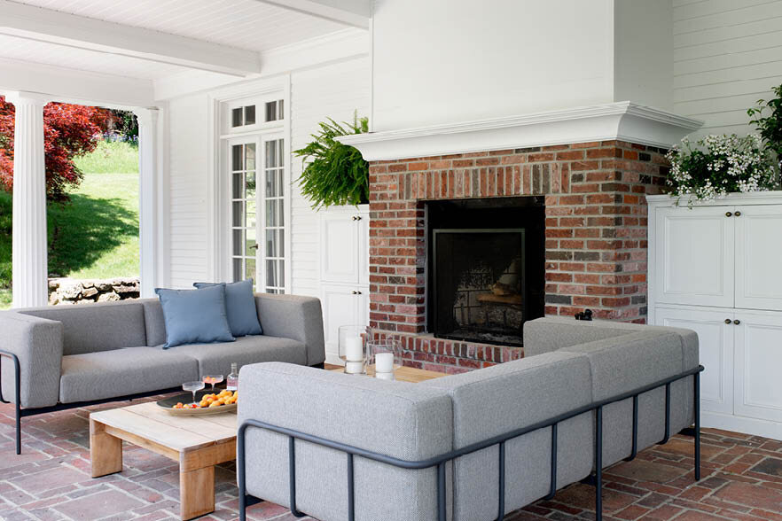 Angled view of the covered porch featuring modern couches with a teak coffee table, a wood-burning fireplace, and greenery through historic columns. As seen on Boston Home Magazine's Instagram.