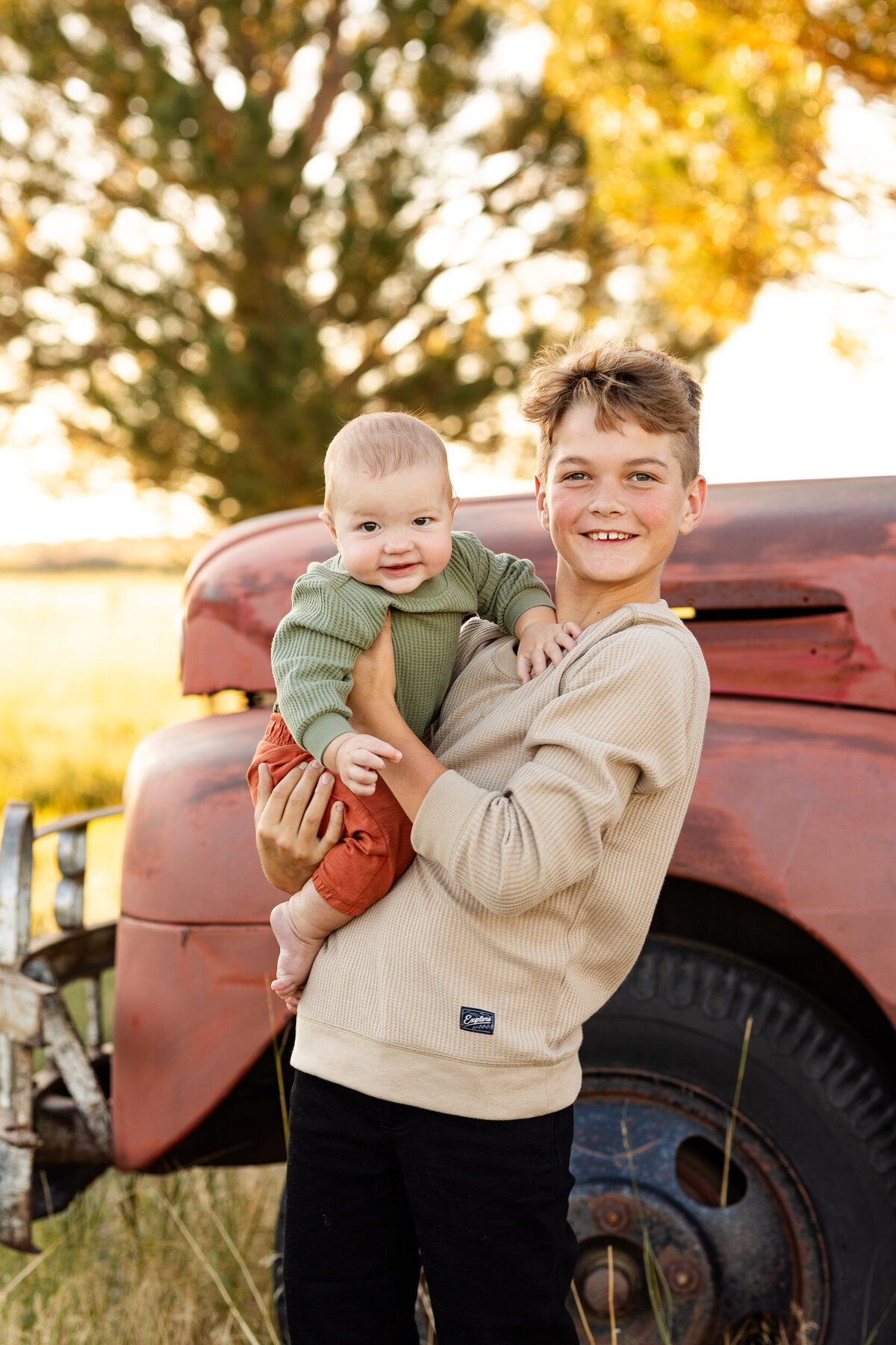 Pre-teen boy holds his baby brother and they smile at the camera.