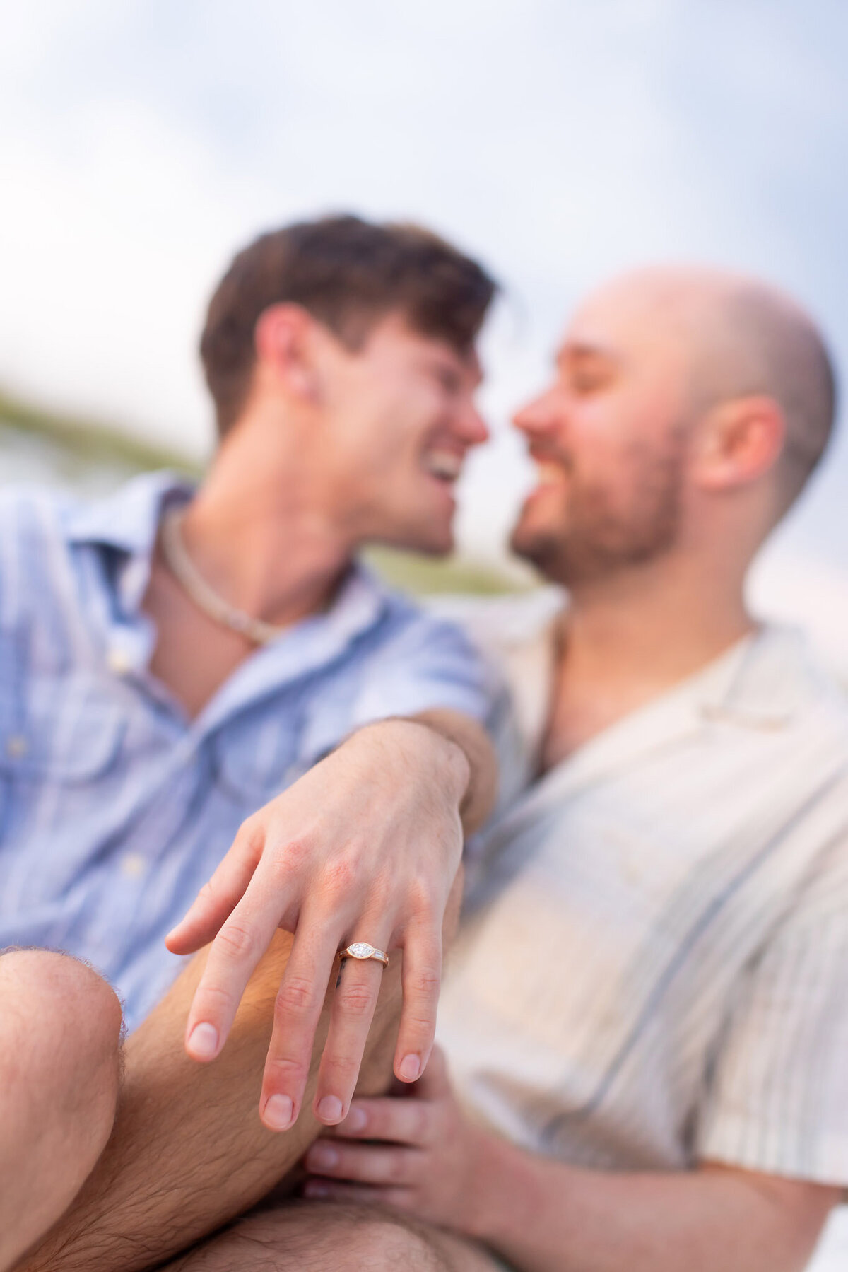 Happy LGBTQ couple sitting in sand smiling with ring visible.