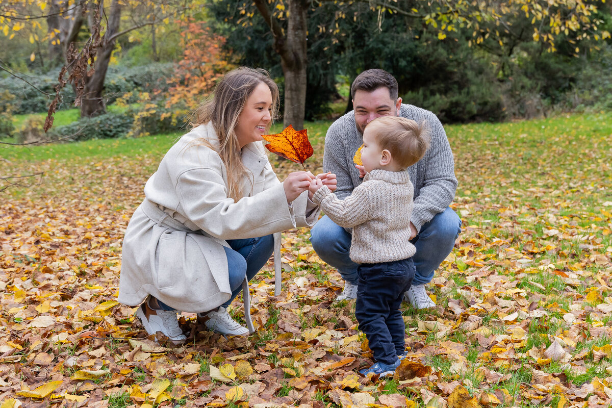 A young family enjoys autumn outdoors, with parents smiling at their toddler holding a leaf in a park covered with fallen leaves.