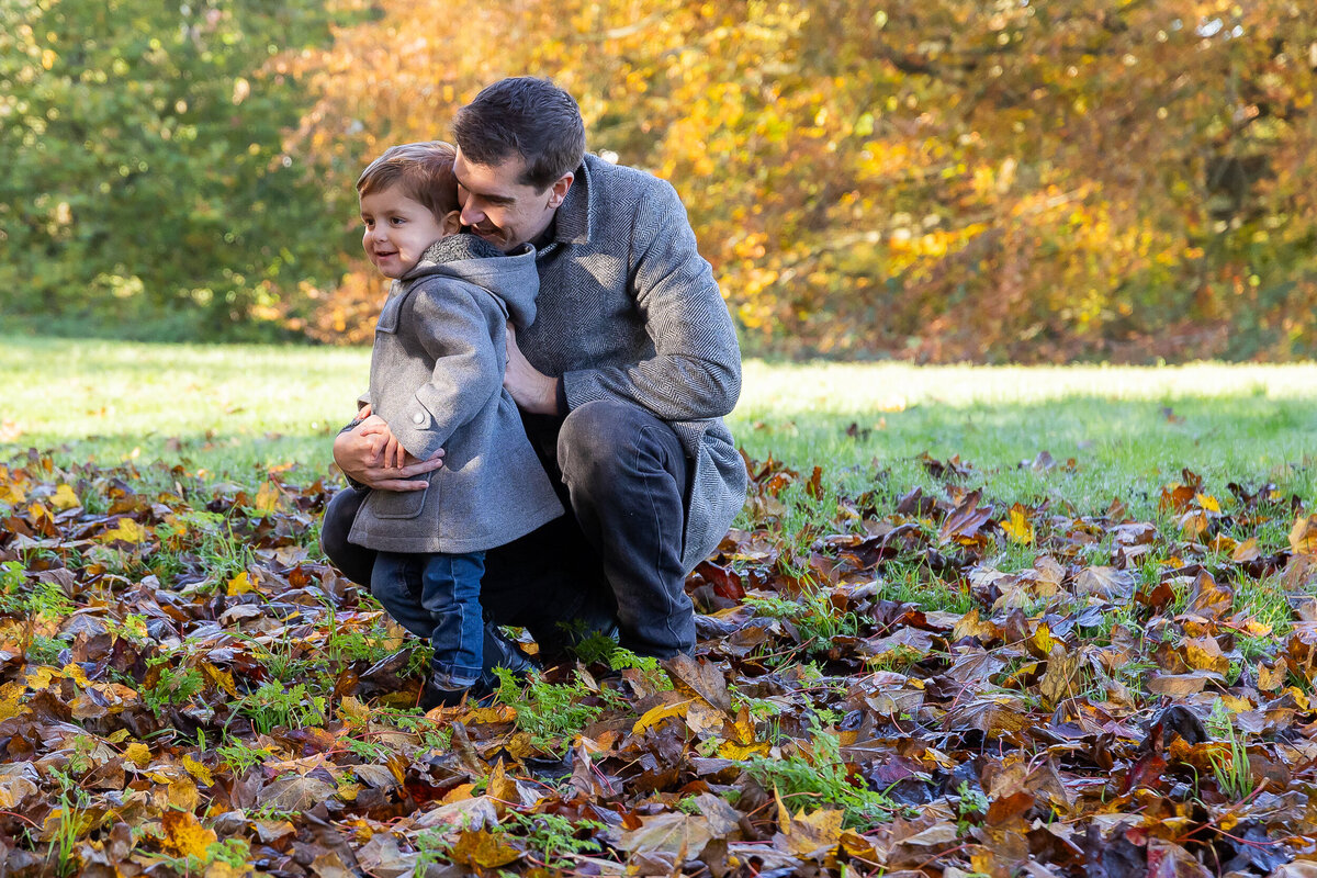 A man and a young boy embracing joyfully while kneeling on fallen leaves in a park during autumn.