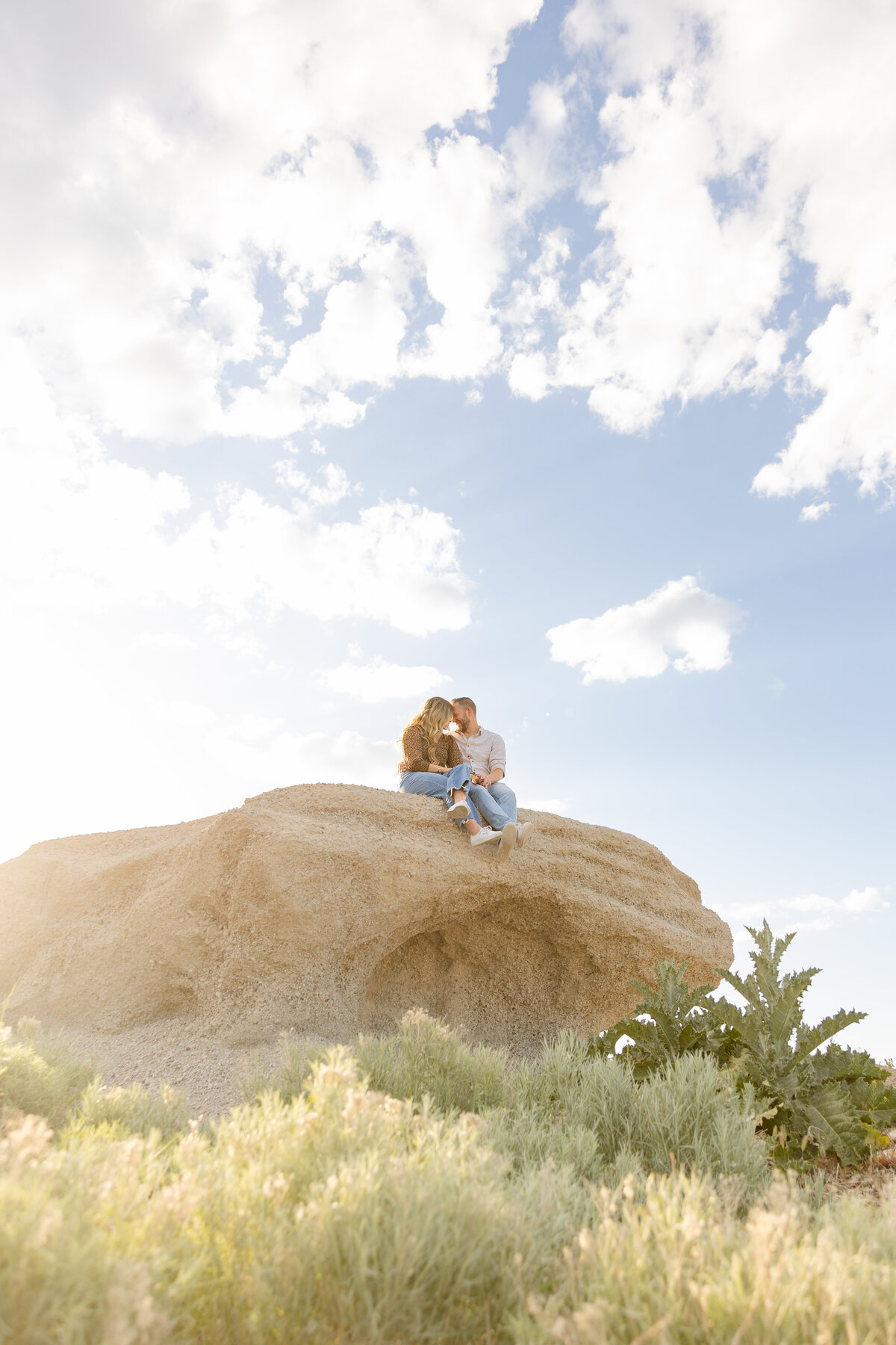 A couple sits atop a large rock formation in Saratoga Springs, Utah, sharing a tender moment under a bright blue sky with scattered clouds. The golden sunlight highlights the natural beauty of the setting.