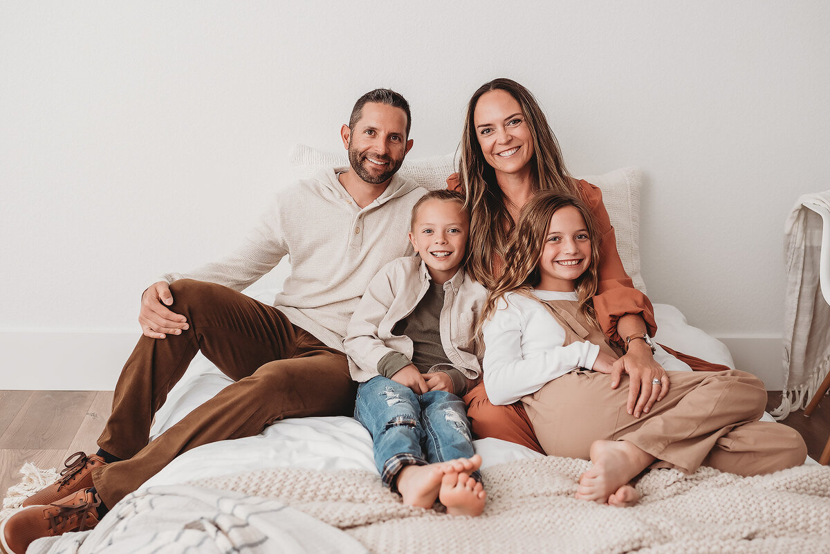 family of four with older kids sitting on bed together looking at photographer's camera
