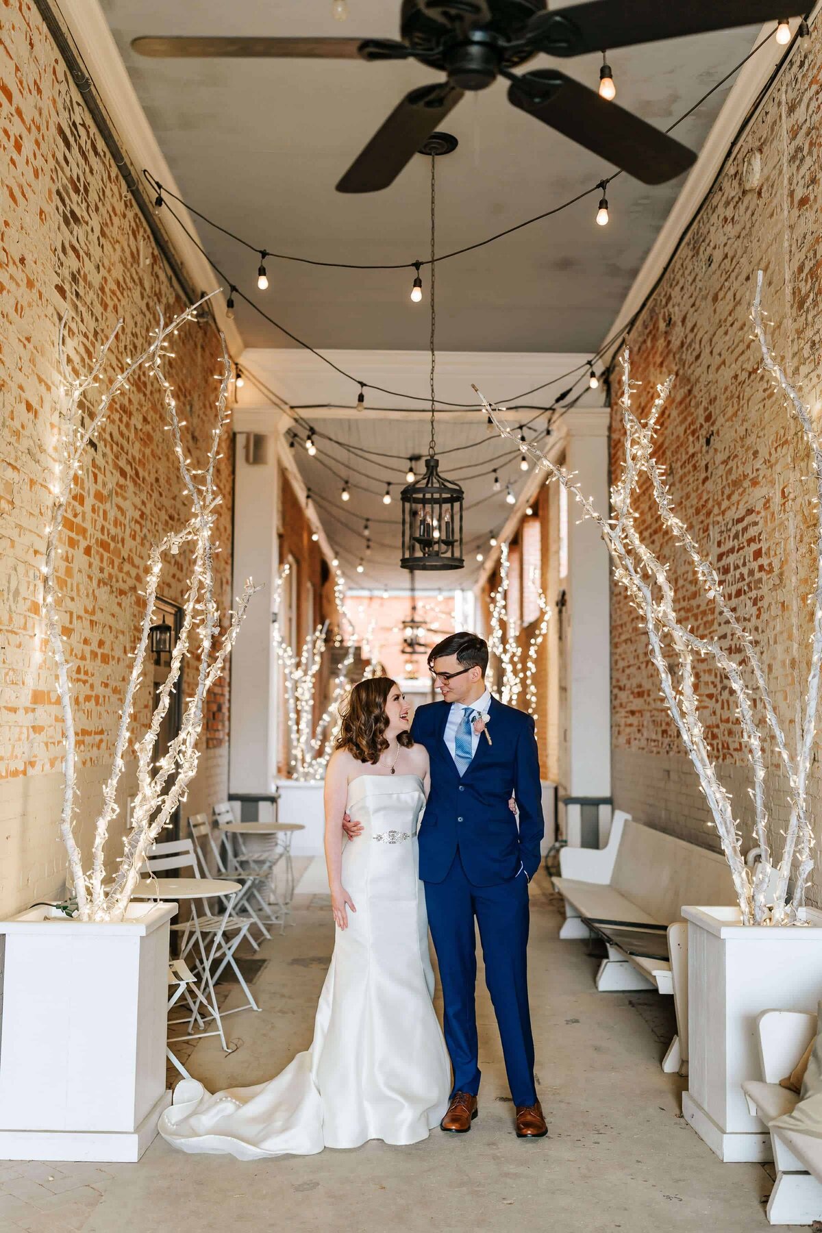 A bride in a white gown and a groom in a blue suit stand together, smiling at each other, in a brick hallway decorated with white lights and bare, illuminated branches.