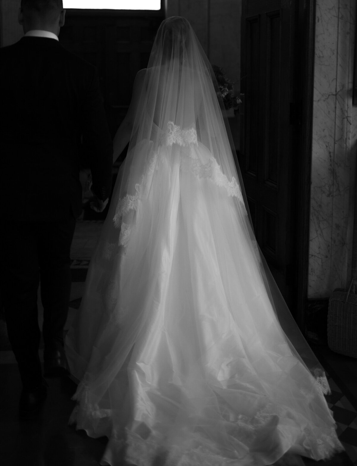 A bride's long lace cathedral veil and gown are lit in a dark hallway in St. Peter and Paul, Detroit