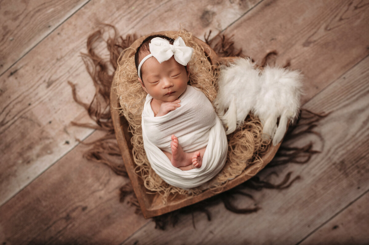sleeping newborn baby girl swaddled in white with her feet and hands poking out posed in a brown wooden bowl next to a pair of angel wings with a fringe brown layer and  wood floor as the background