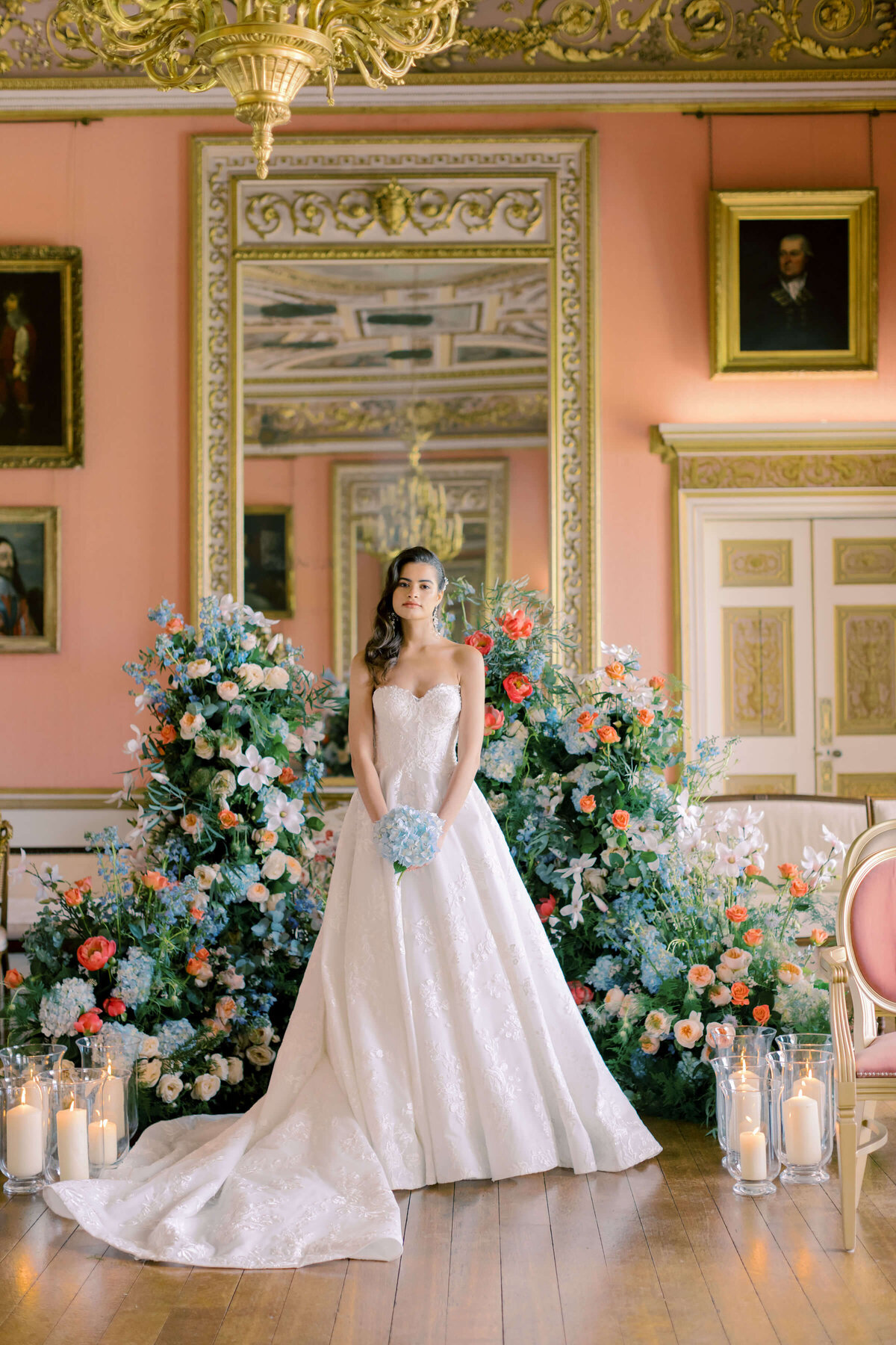 bride holding a single blue hydrangea standing in front of a flower wall of blue and coral flowers with candles at the base in avington park’s ballroom