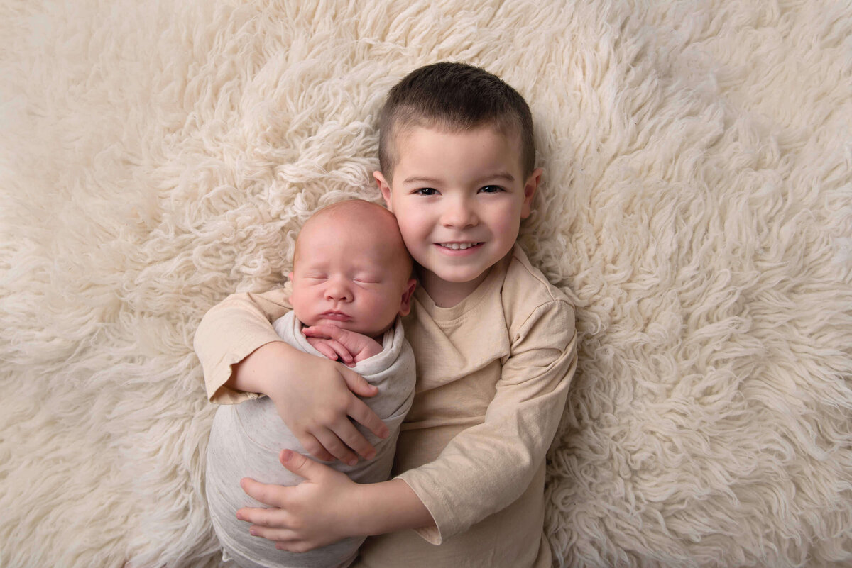 big brother holding newborn baby boy brother on a cream coloured rug for their Burlington, Ontario newborn photography session.
