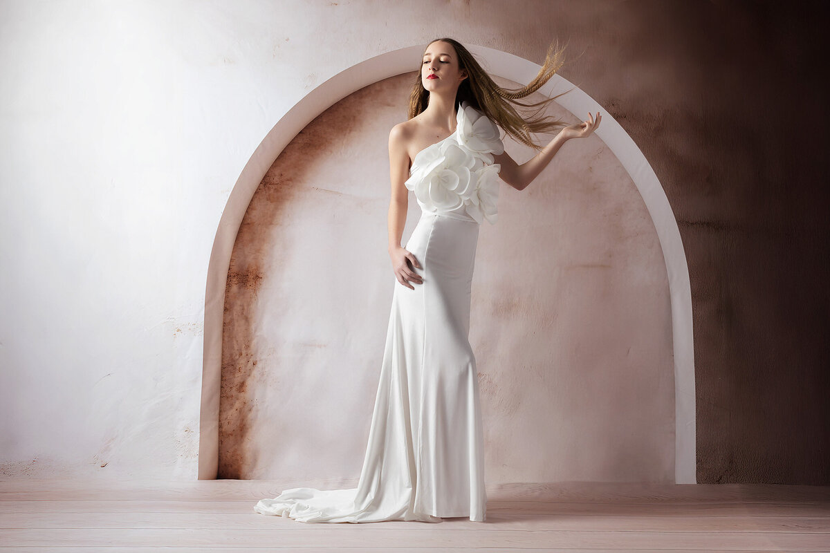 A model wearing a white, form-fitting gown adorned with large white flowers on one shoulder stands in front of a soft, curved blush-toned background. Her hair is caught in motion, creating a dynamic and elegant scene for a Dallas senior photographer session.