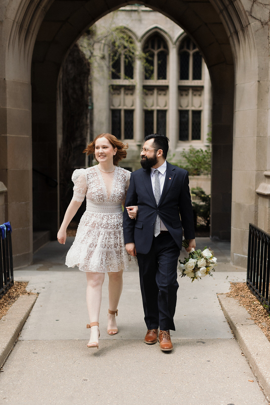 Happy couple walks quickly outside of church, They're smiling and their arms are linked, groom carries the bouquet and looks at bride.