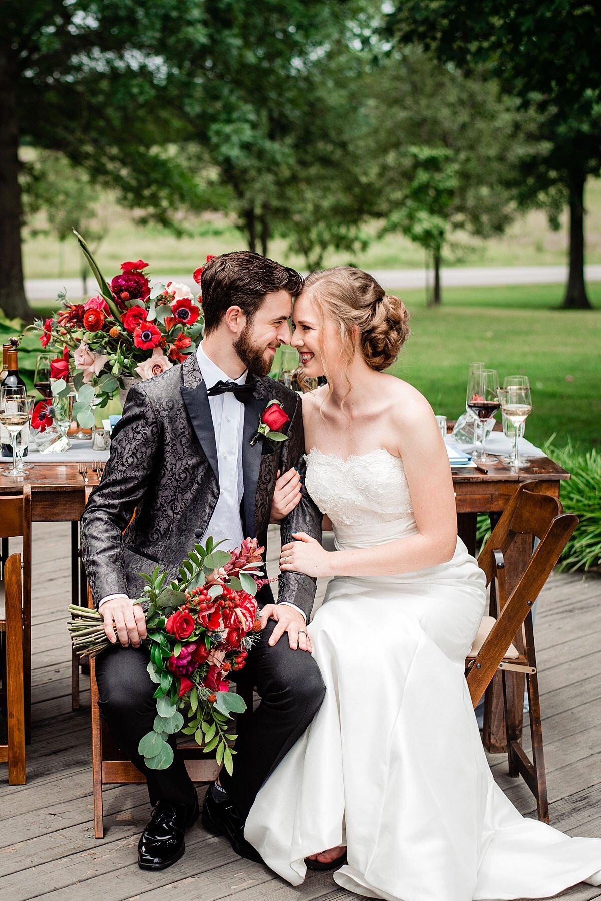 The bride, dressed in a white silk wedding dress and a strapless lace bodice with a  sweetheart neckline embraces the groom who is wearing a black tuxedo with a black silk tuxedo jacket with an embroidered paisley design and red rose boutonniere. The bride and groom, touching foreheads, sit in front of a dark wood farm table  decorated with a large red and blush floral centerpiece. The groom holds the cascading bridal bouquet of red ranunculus, hypericum berries, blush roses, red anthurium, red roses, red ranunculus, red anemones, red proteas, blue thistle, red dianthus and assorted cascading greenery.