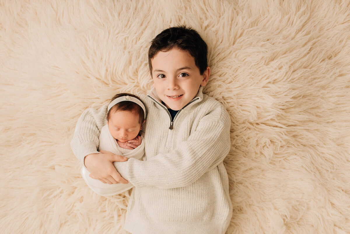 A close-up of a new big brother on a white rug with his newborn baby sister.