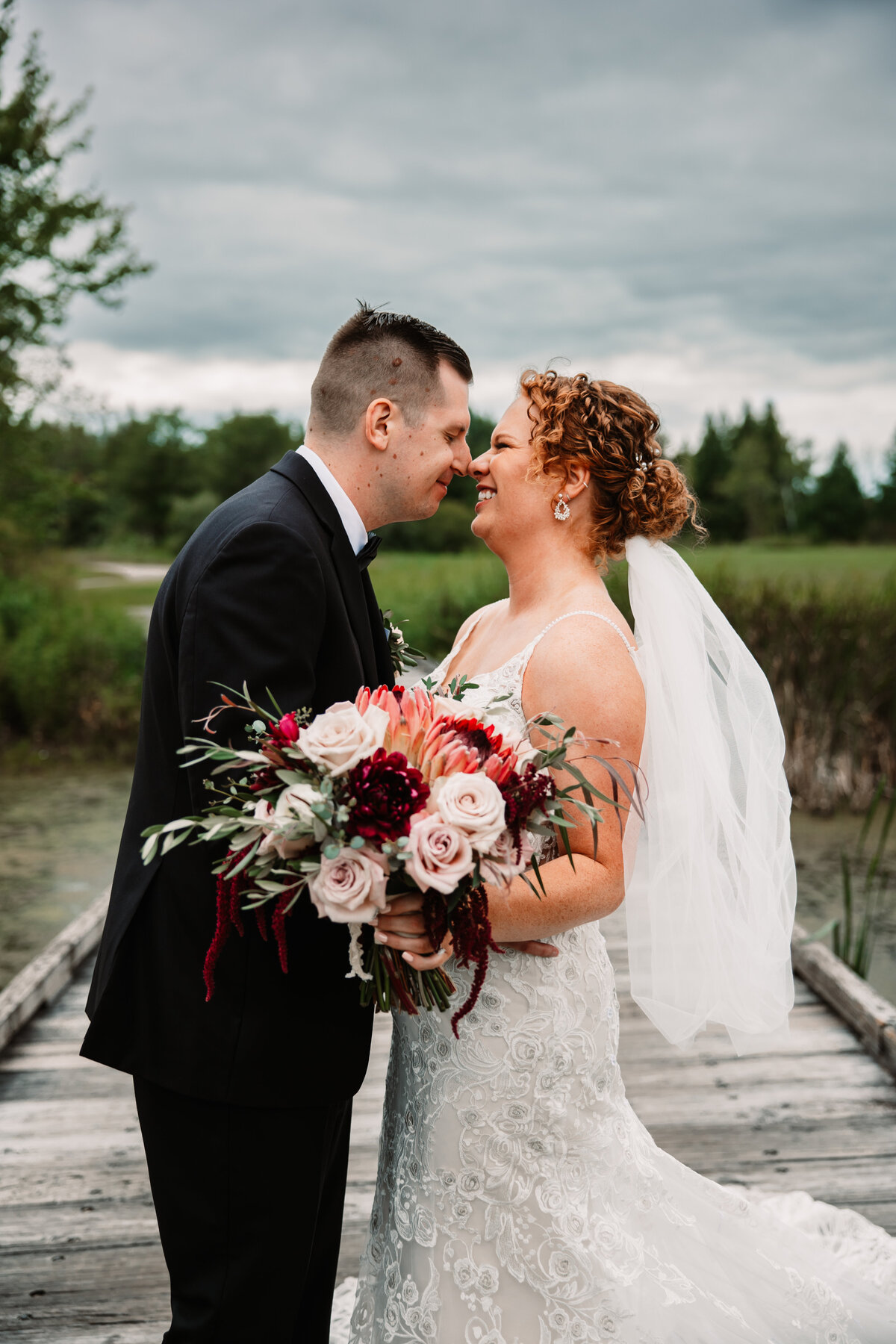 A bride and groom touch noses while giggling and standing on a wooden bridge