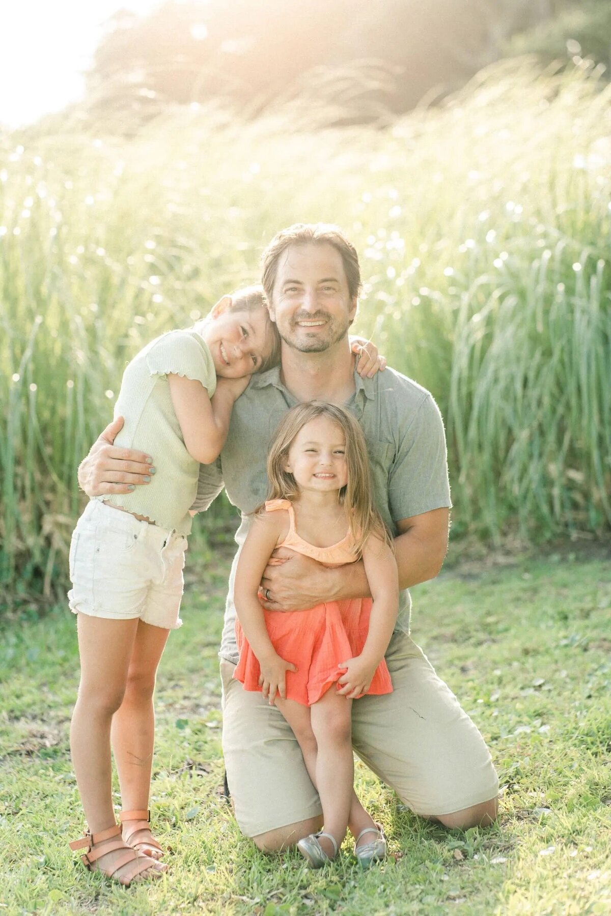 A dad kneeling on the ground with one child resting their head on his shoulder and the other leaning in front of him.