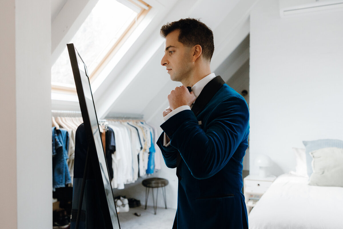 Groom in blue velvet suit fixing his bowtie in front of a standing mirror.