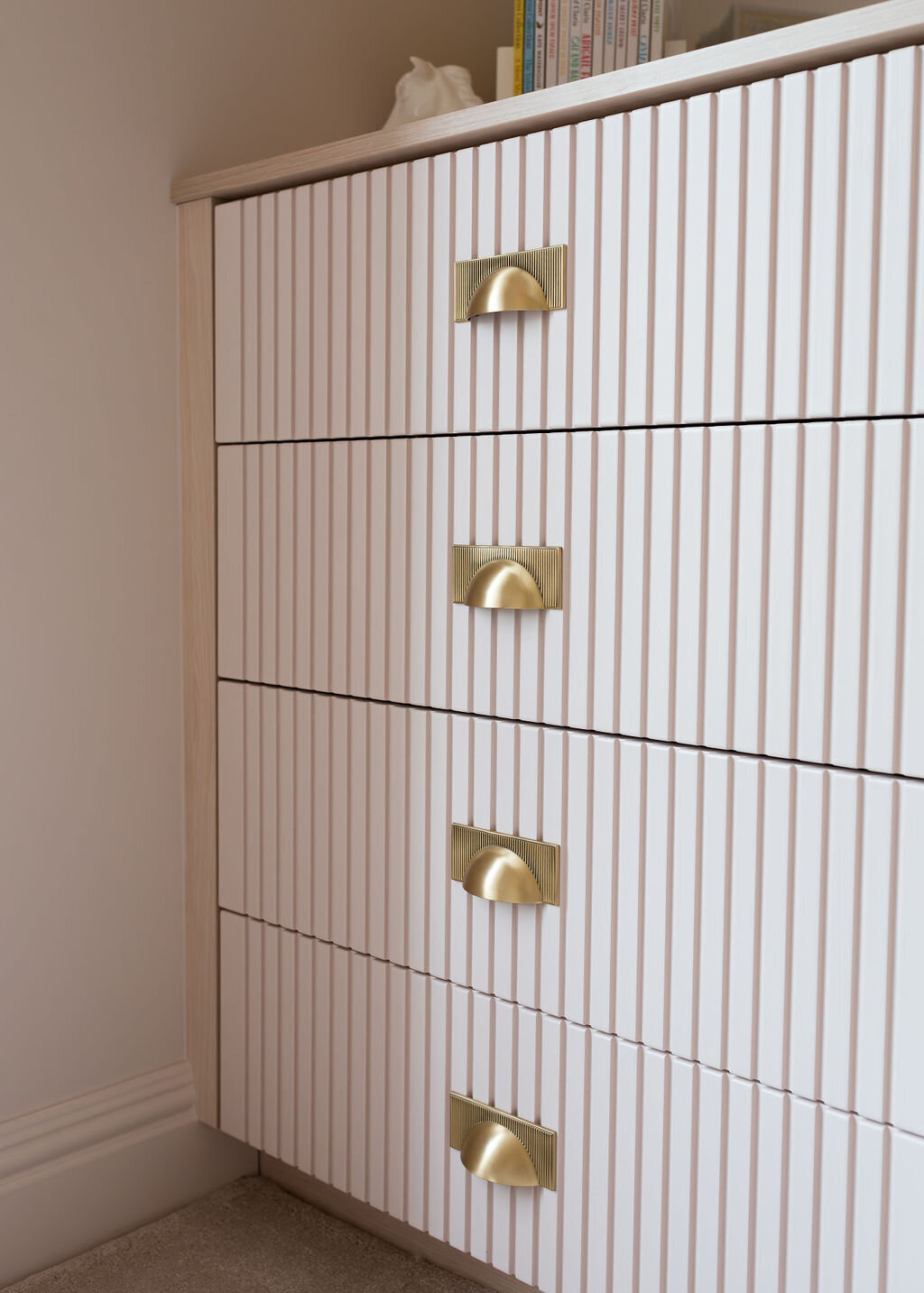 A close-up view of a modern pale pink dresser with gold half-moon handles. The dresser has a ribbed texture on the drawer fronts and features four drawers.