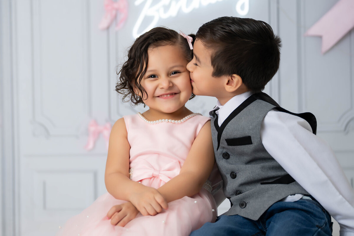 Little girl in a pink dress sitting next to a boy in a suit vest who is kissing her on the cheek.