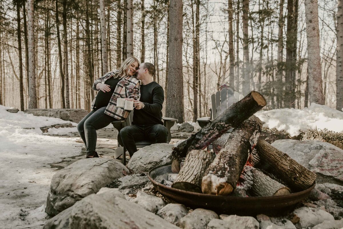 Expectant mom and dad in Exeter, Ontario forest for maternity photoshoot. An  unlit campfire is in the foreground and mom and dad are cuddling on a chair in the background.