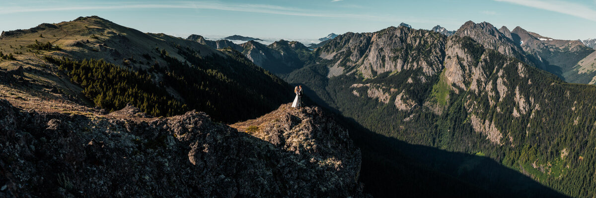 a panoramic image of sweeping mountains at sunrise. A couple, in their wedding attire embraces on a peak as their adventure elopement photographers capture the moment. 