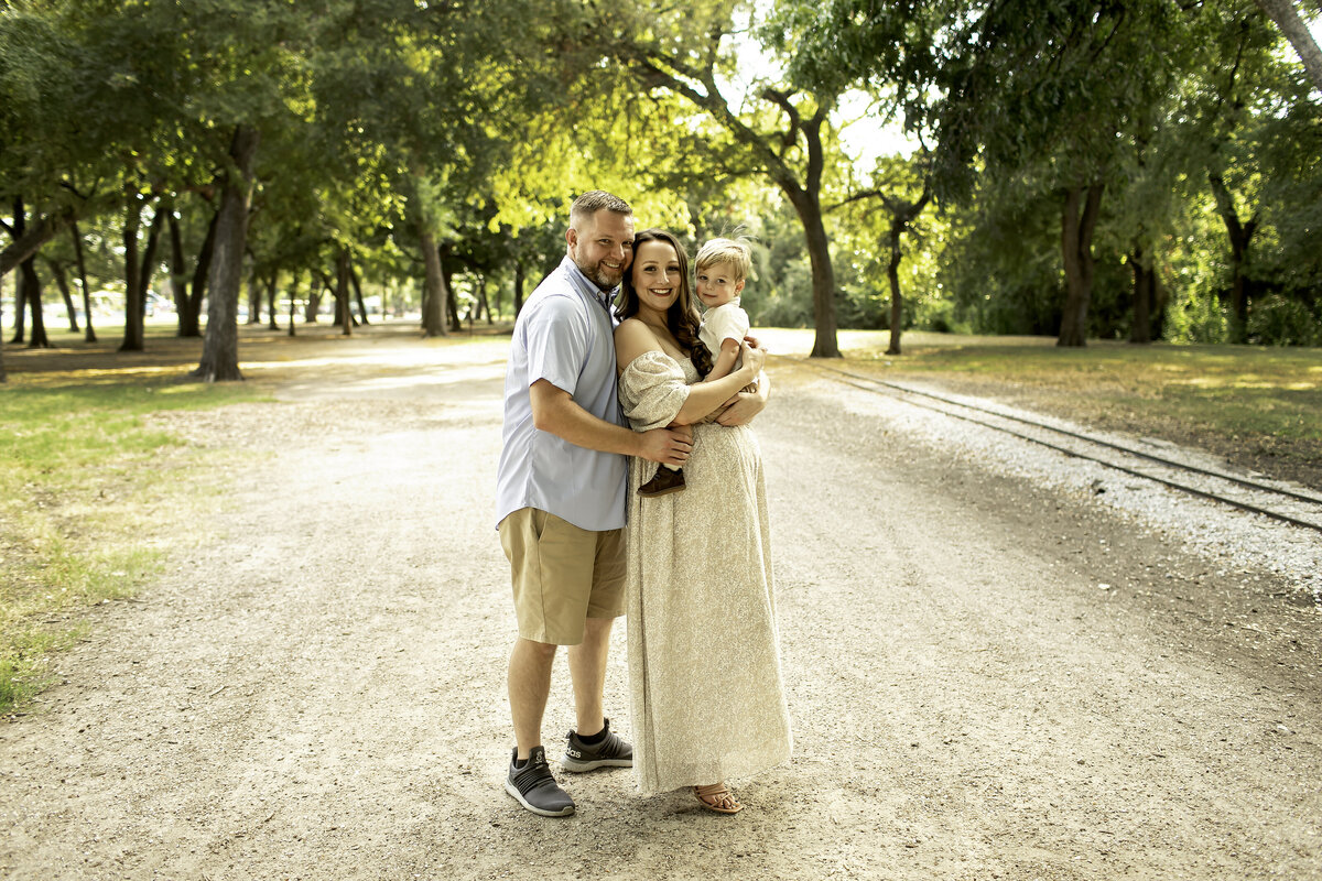 A romantic maternity photo of mom and dad standing in the park, with mom gently cradling her belly while dad wraps his arm around her, captured by Chunky Monkey Photography.