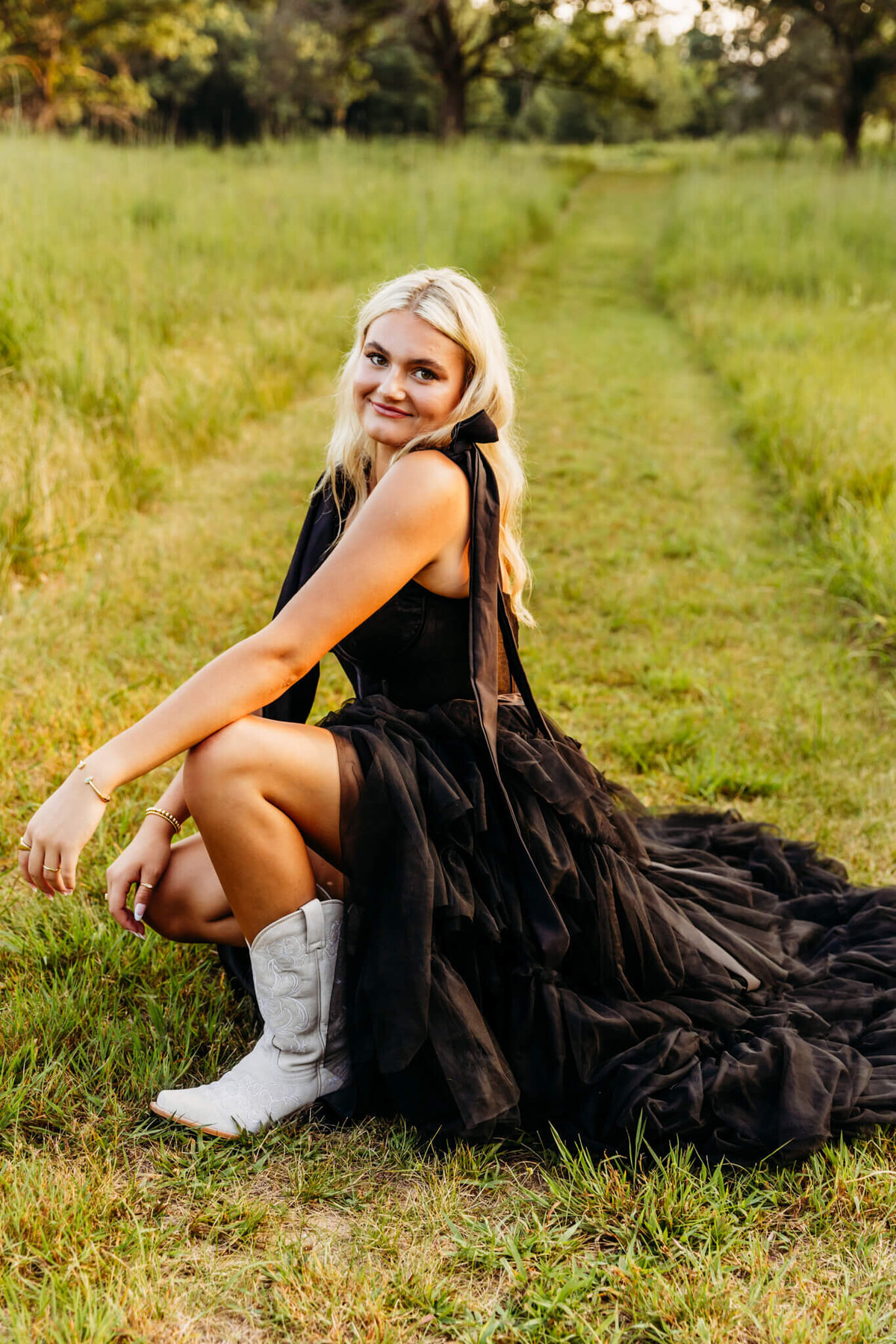 blonde teen girl kneeling on a grassy path in a black dress with one white boot sticking out for her senior photos.