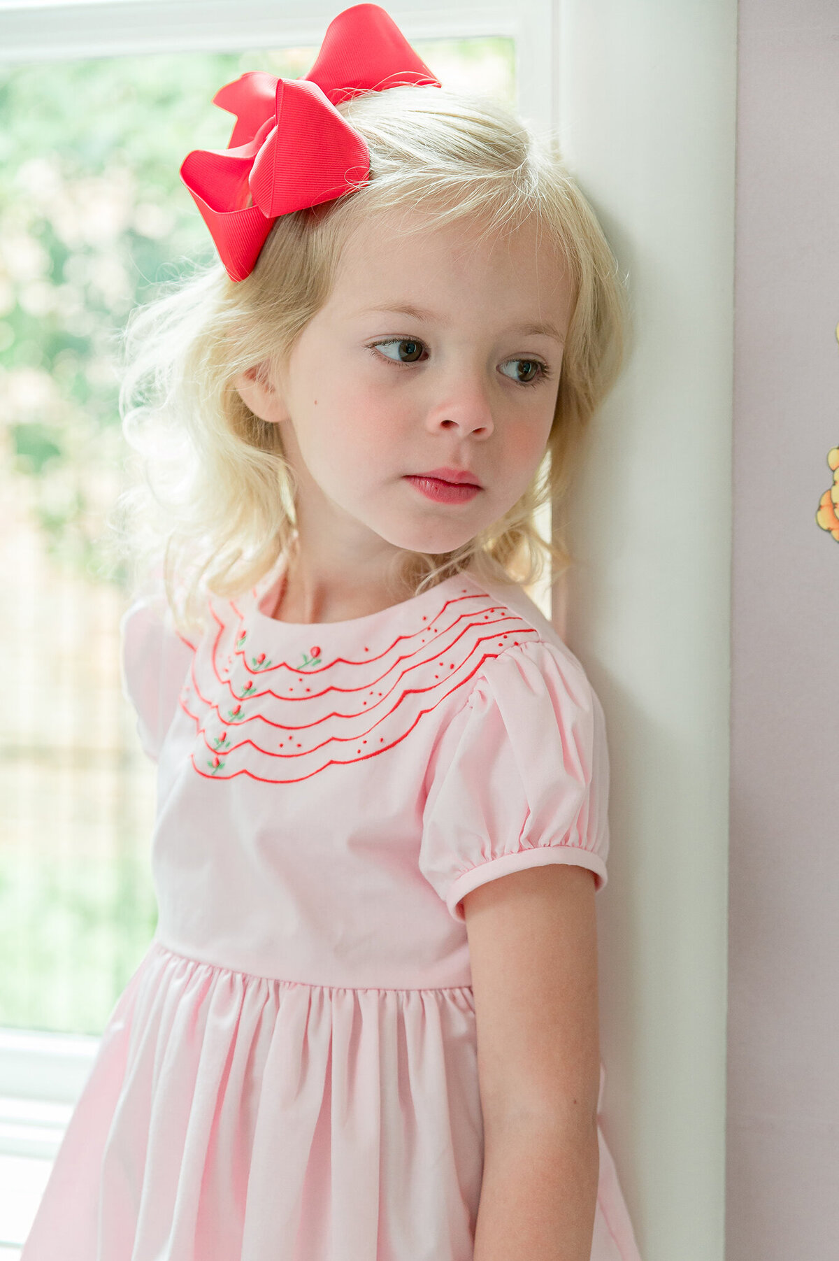 Blonde preschooler leaning up against a window frame gazing into the distance.