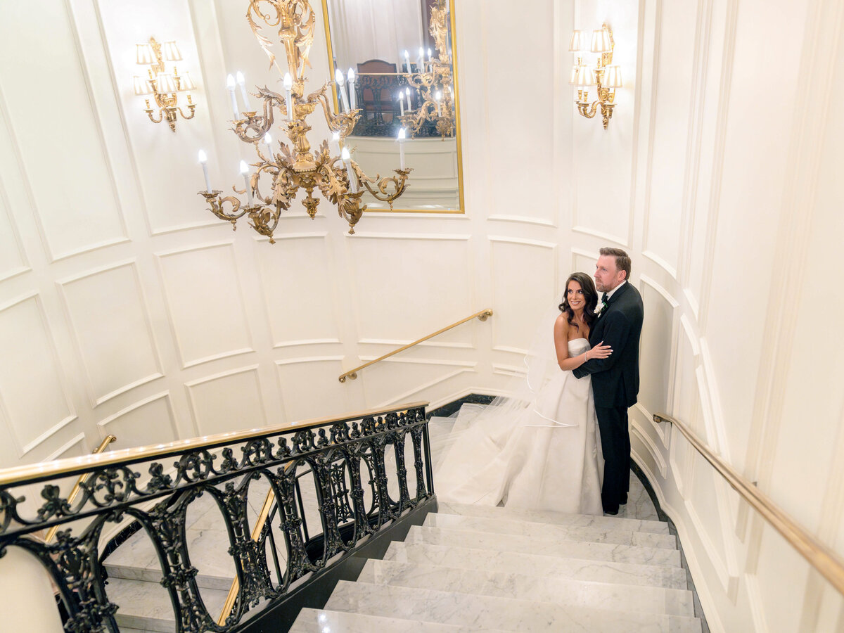 A couple poses on a grand marble staircase beneath an ornate chandelier. The woman is in a white wedding dress and the man is in a black tuxedo. The elegant setting features white paneled walls and a large mirror.