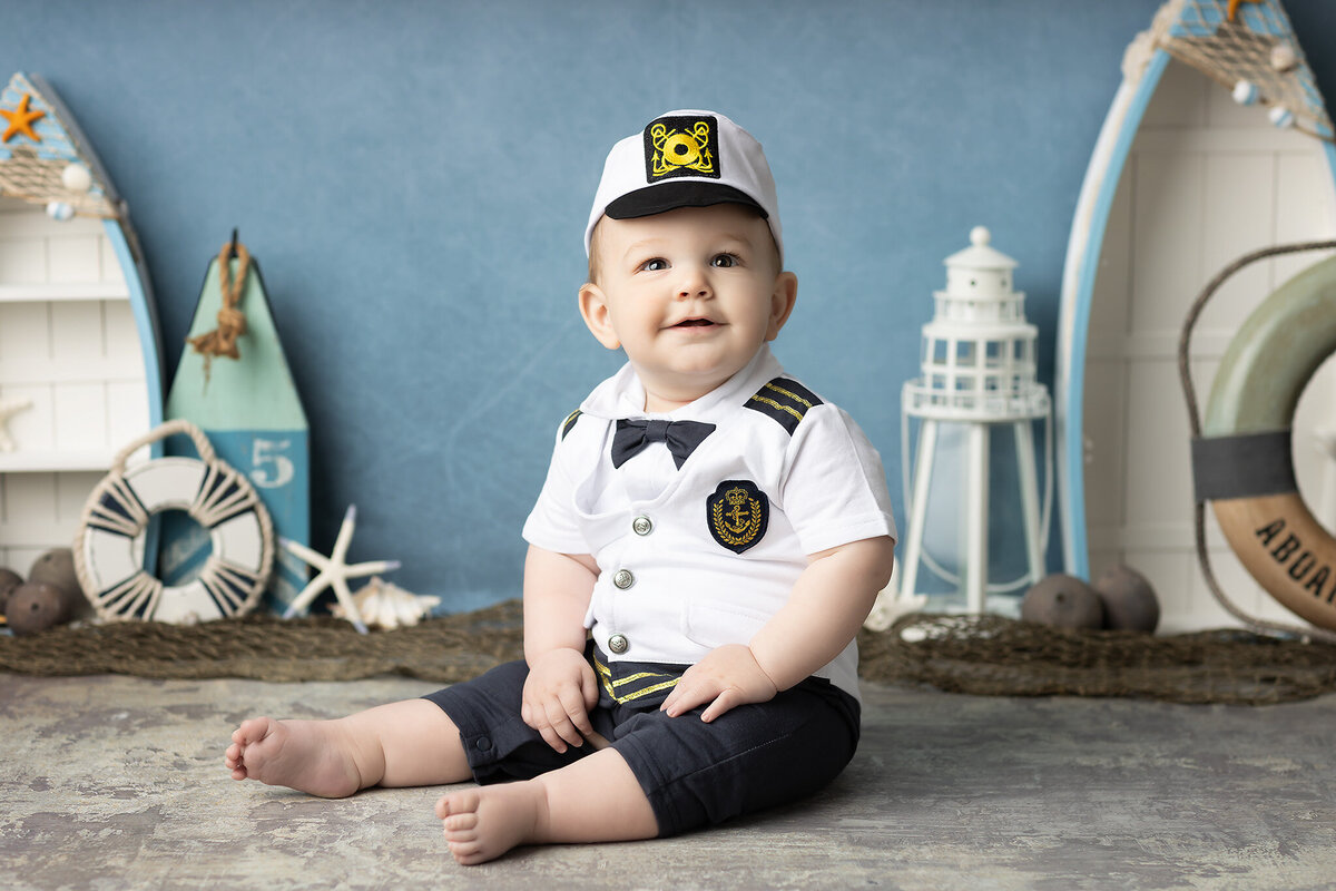Smiling baby boy dressed as a sailor in a navy-themed milestone photoshoot with lighthouse and life preserver props by a Dallas milestone photographer.