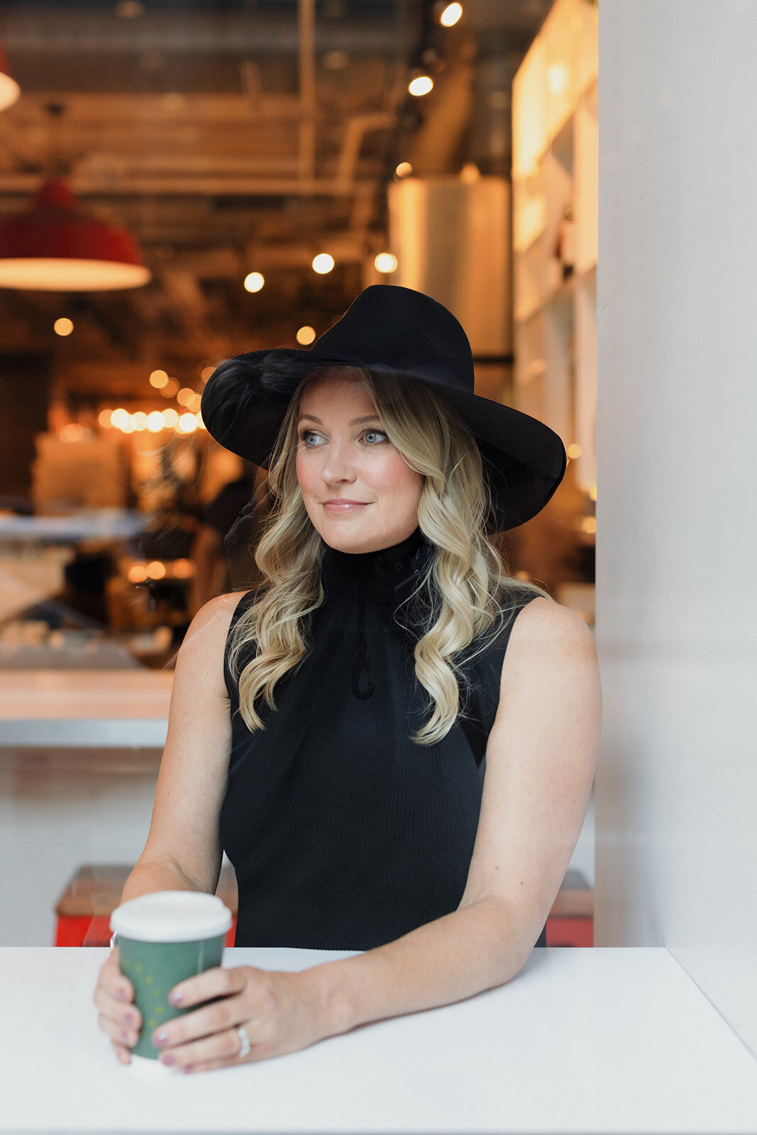 blonde woman in black sleeveless top and black fedora holding green cup of tea. She looks out window, photo is shot through glass so there are some street reflections in glass.