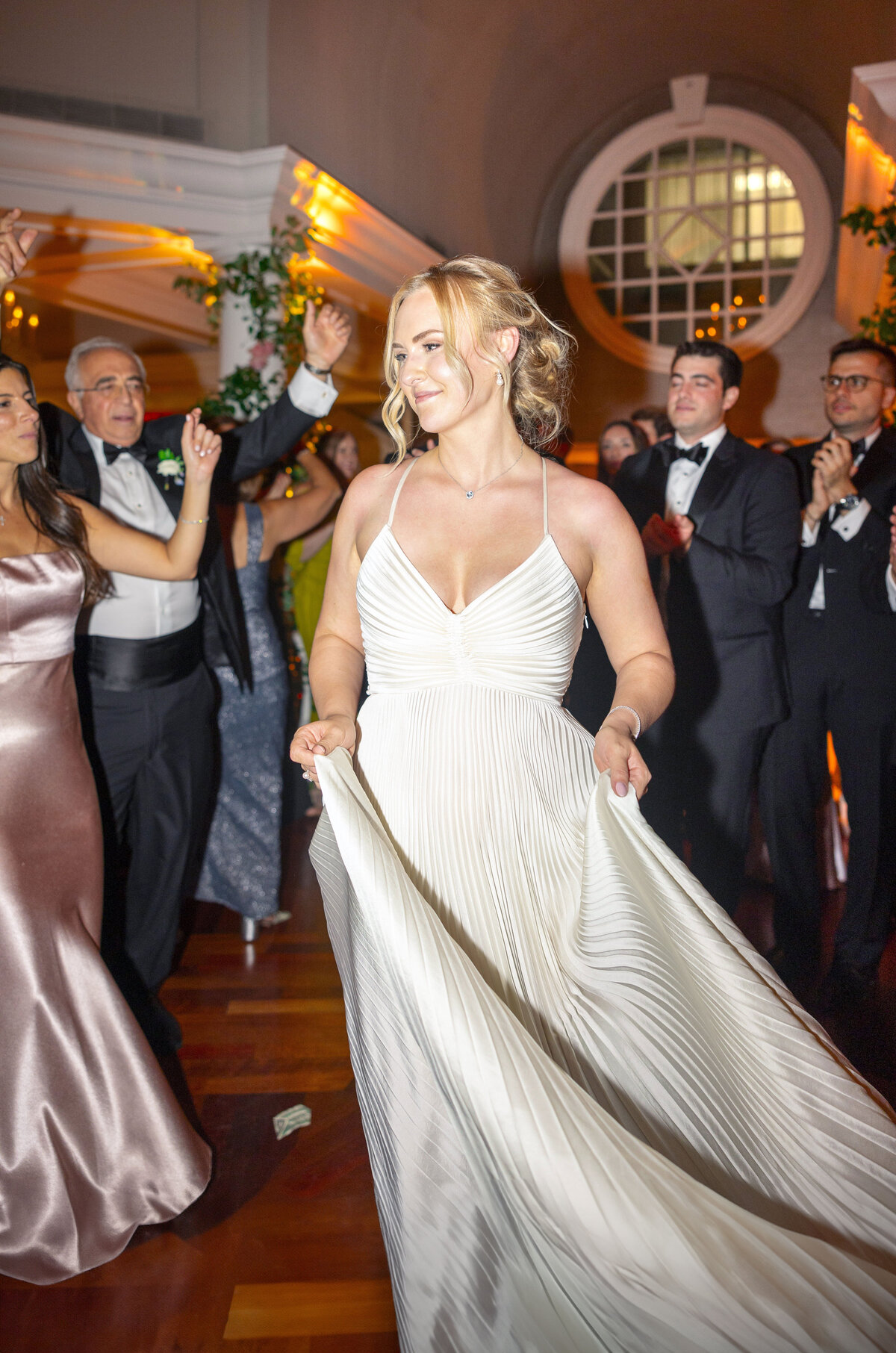 A woman in a white gown dances at a formal event, surrounded by cheering, well-dressed guests. The setting is elegant, featuring warm lighting and decorative vines. A circular window is visible in the background.