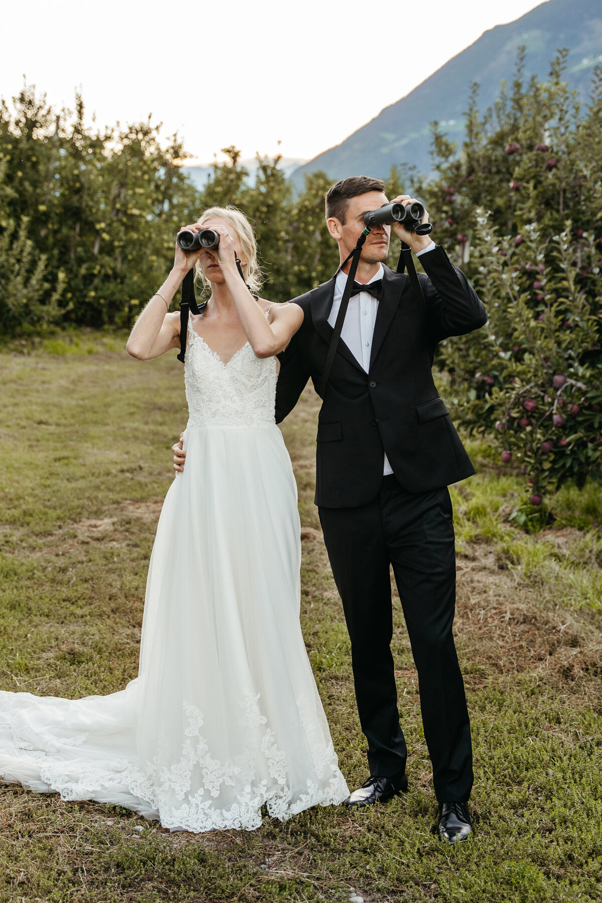 Bride and groom birdwatching with binoculars in orchard