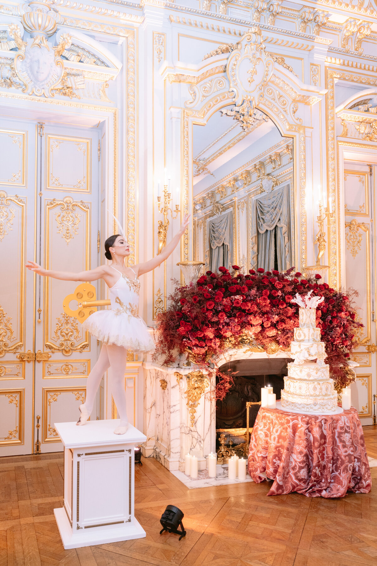 A ballerina in a white tutu stands on a pedestal with arms outstretched in a lavishly decorated room. Behind her, there's a grand fireplace adorned with an arrangement of red flowers and a tall, ornate white cake with multiple tiers. The room features ornate gold accents.
