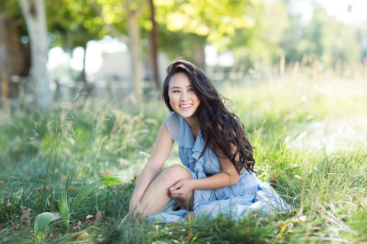 Casual senior portrait in a grassy park setting, with the teen sitting cross-legged and smiling at the camera.
