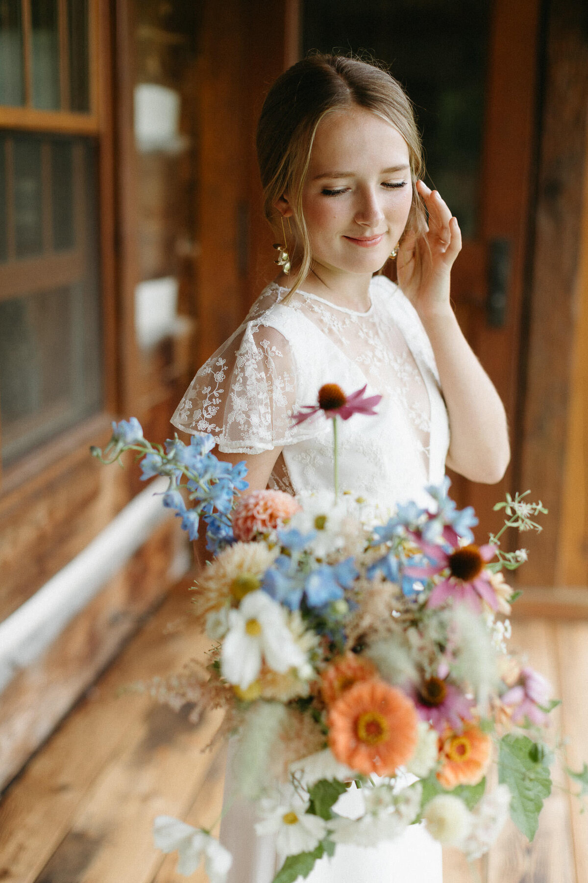 A woman in a white lace dress stands on a wooden porch at her private estate wedding, gently touching her hair as she looks down at her colorful bouquet. This serene moment is beautifully captured by Toronto wedding photographer, Jennifer van Son.