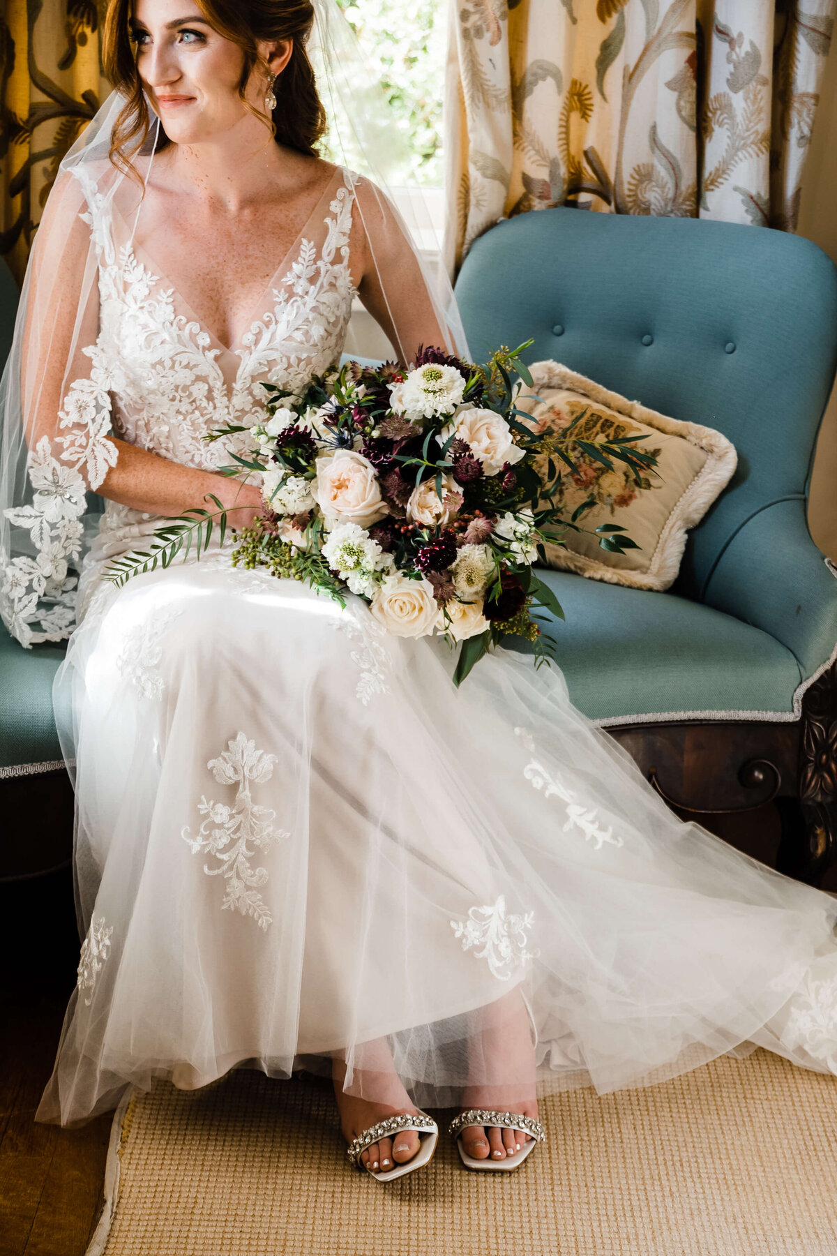 wedding portrait of bride sitting on a vintage couch in a old house that has been converted to a wedding venue holding her floral bouquet on her lap and gently smiling as she looks out a window onto the garden