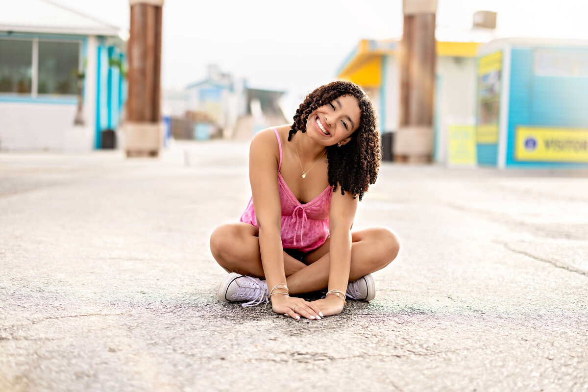 Winter Garden senior session at Cocoa Beach with a girl sitting criss-cross on the ground, hands down, smiling with her head tilted softly.