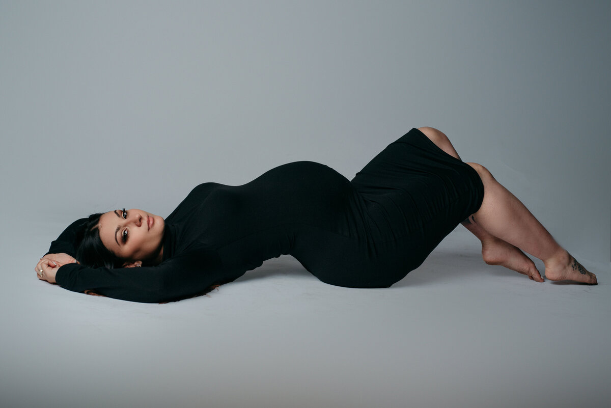 34 week pregnant woman posed laying on her back in a black knee length dress on white background looking at camera with closed mouth smile