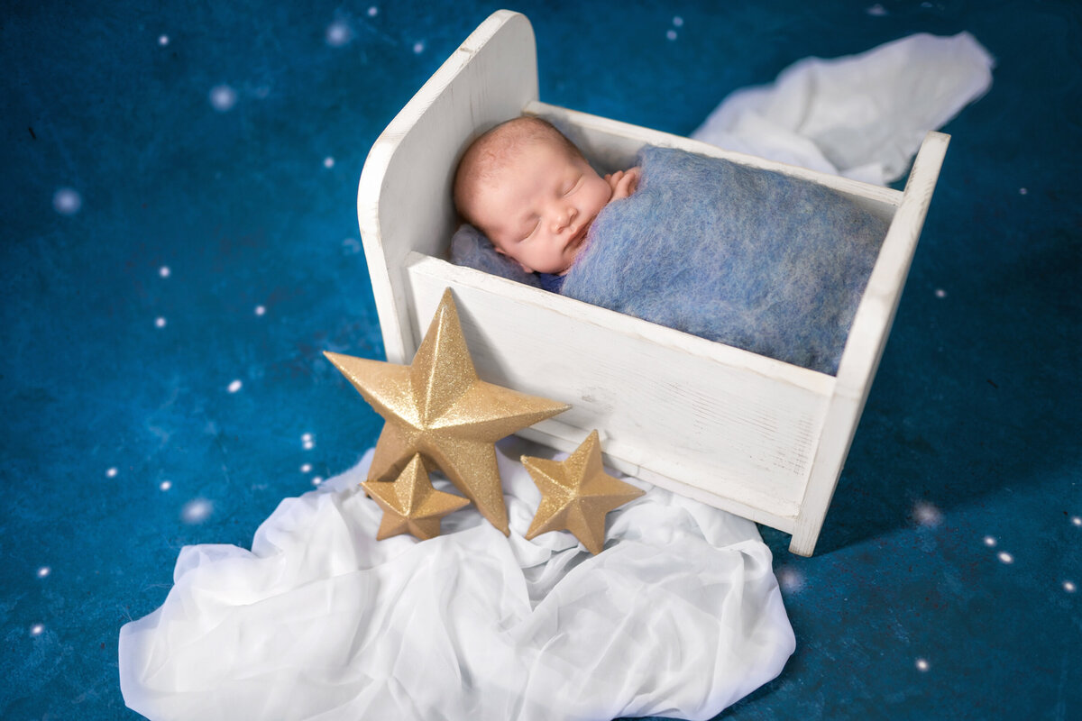 Baby boy under a blue blanket in a tiny white bed.  He is on a blue background that looks like the sky.  There is a white wrap like clouds and gold stars.  Baby is asleep.