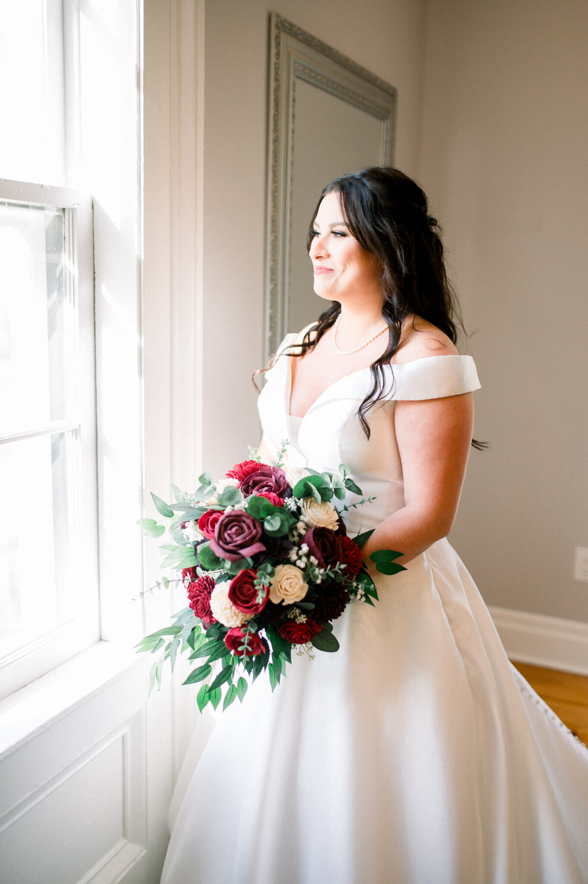 Bride standing in front of window  holding red bouquet. Captured by Niagara wedding photographer