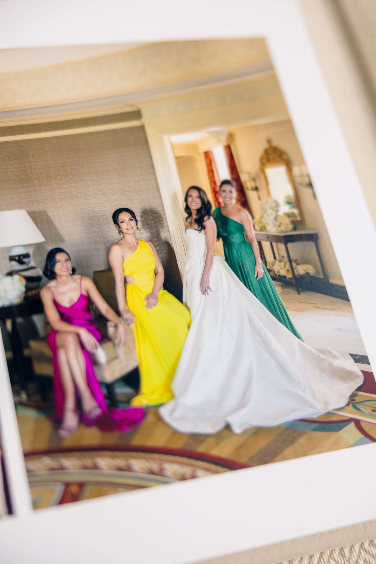 Four women pose elegantly in a room. One wears a white gown, and the others are in vibrant dresses: pink, yellow, and green. A mirror and decor can be seen in the background.