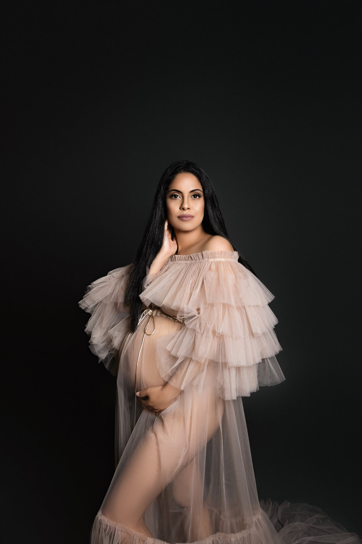 Maternity photos captured by top London, ON Maternity Photographer Amy Perrin-Ogg. Woman in tulle gown is standing side profile to the camera looking over her shoulder and holding her bump.