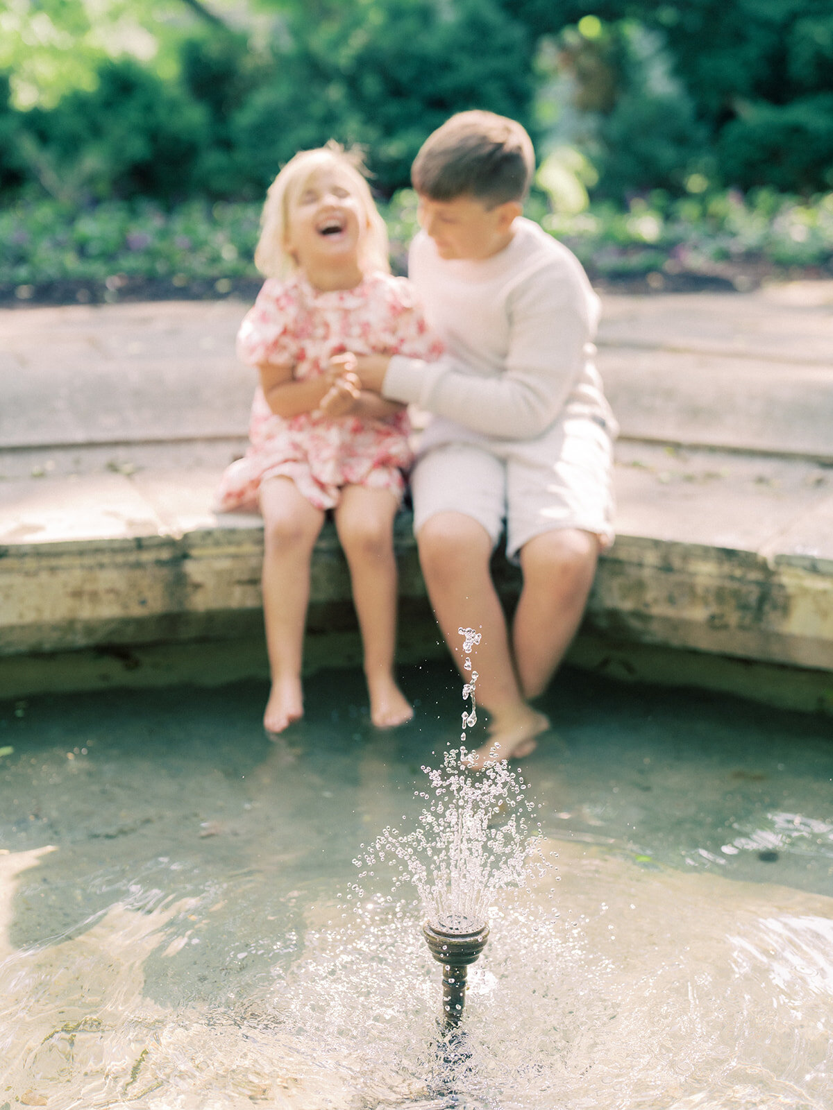 Brother and sister sit on edge of fountain with feet dipping in, laughing.